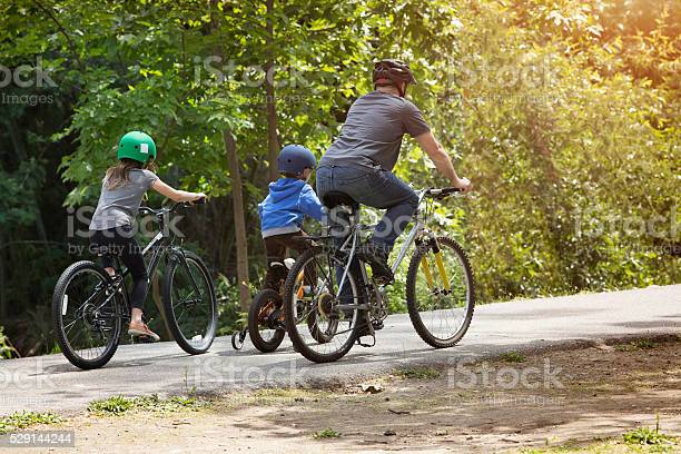 Rear view of a Father leading his kids on bike ride in park. they are all wearing helmets.