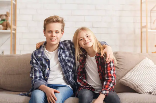 Cute Brother And Sister Embracing Smiling To Camera Sitting On Couch At Home. Siblings Friendship Concept